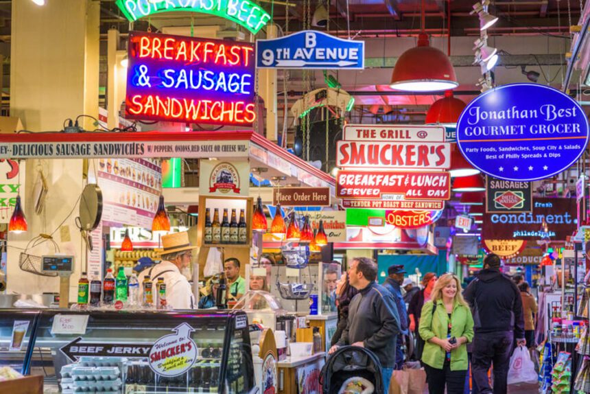 Neon signs light up over stalls inside of the historic Reading Terminal Market. Shoppers are shown in the aisle, workers are behind counters serving customers.