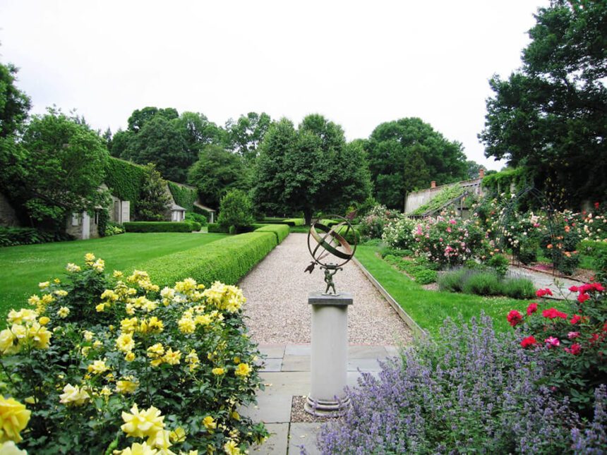 An outdoor garden is filled with lush green lawns and colorful flowers. A large bush with yellow flowers is shown in the bottom left-hand corner. To the right, there are beautiful purple flowers and red roses blooming. Along the left side, there are bushes with pink flowers blossoming. A sculpture is in the center.