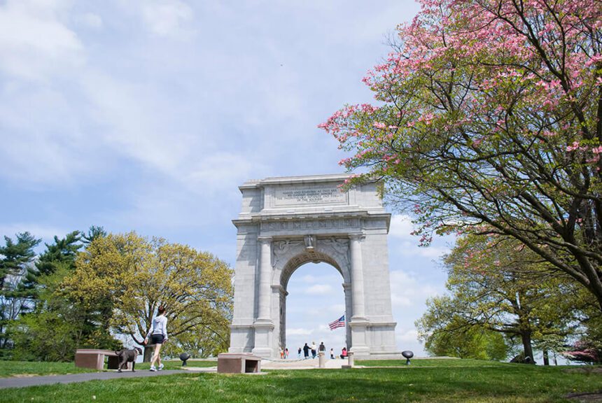 A large white arch stands tall in the middle of a park. To the left, a woman walks her dog on a trail. To the right, there is a large tree with pink blossoms. Through the arch in the distance, there are numerous people shown walking around.