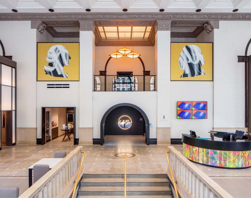 An open lobby is shown. There is a colorful front desk shown to the right of a staircase. There are large paintings hung on either side of the space. There are large light fixtures hanging from the ceiling. 