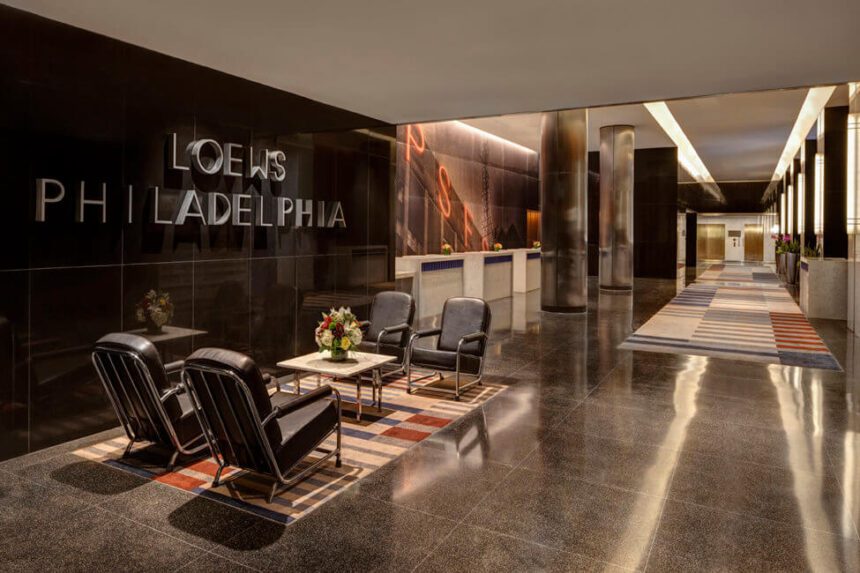 There are four chairs placed around a small table with flowers on it. The wall to the left reads LOEWS PHILADELPHIA. To the right, a long hallway is shown with a carpet with a gray, blue, and red pattern.
