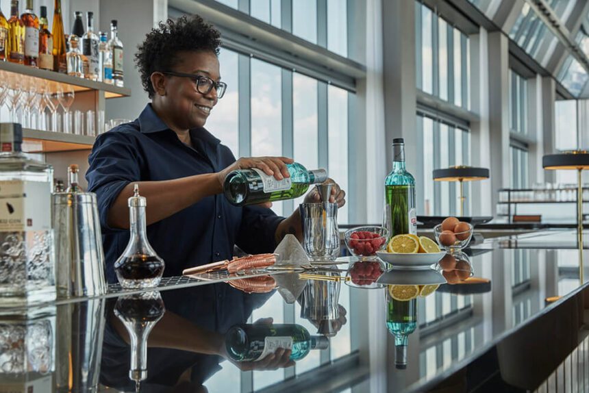 A woman stands behind a bar and prepares a cocktail. she holds a bottle in her right hand and pours its contents into a shot glass shown in her left hand. Bowls of fruits including what appear to be red raspberries, oranges, and lemons appear on the counter. Large windows in the background are letting in natural light. A stocked bar is behind the woman making the drink.