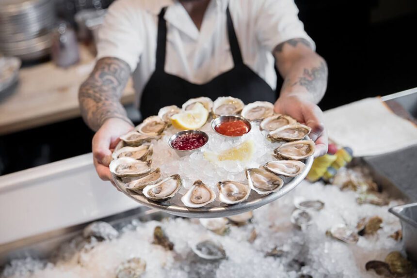 A man holds a platter of shucked oysters and sauces out if front of him. A counter with ice appears underneath.