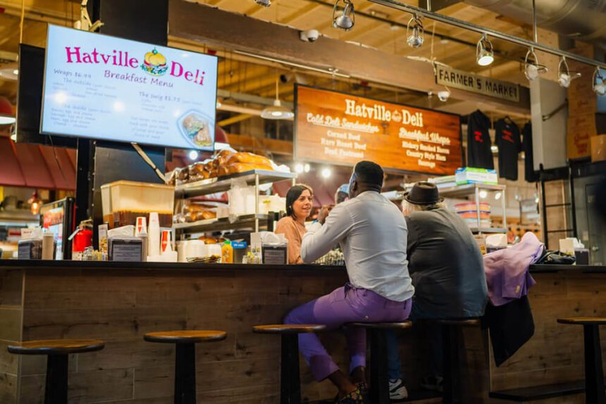 Two men are shown sitting at a counter in front of a stall inside of Reading Terminal Market. Two signs hang overhead. One reads Hatville Deli Breakfast Menu.