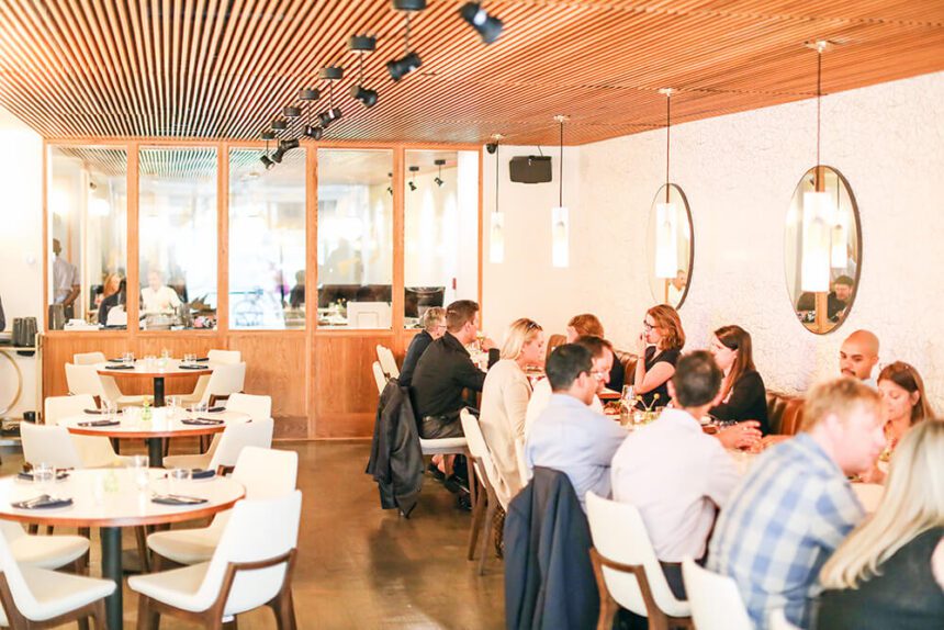 A brightly-lit indoor restaurant space is show. People are seen sitting on the right along benches against the wall and chairs pushed up against tables. There are empty tables set up to the left.