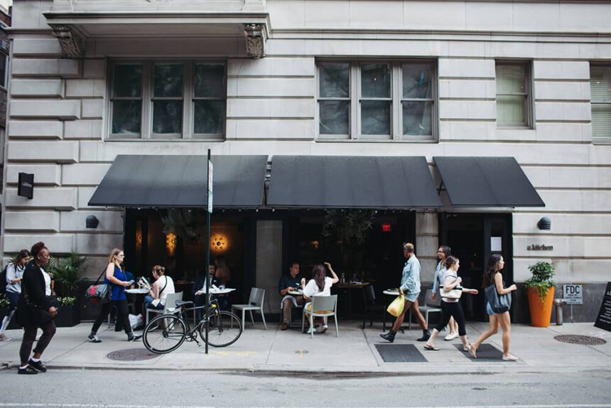 A black awning hangs over several tables situated on the sidewalk. People are shown sitting at the tables as other people walk pas them. To the right, the name of the establishment "a kitchen" is shown written on the side of the building.