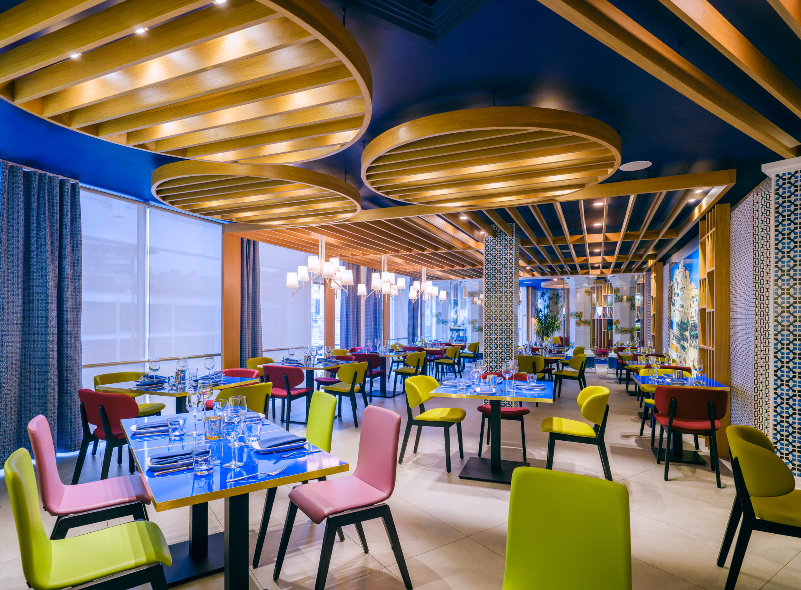 Pink and green brightly colored chairs are seated around shiny blue tables with large lights hanging overhead, large windows are off to the left letting in natural light, as well