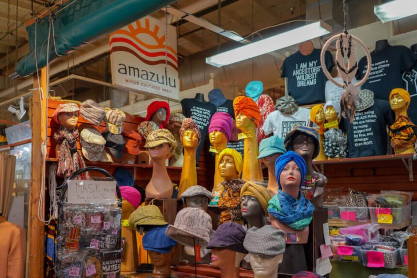 A bunch of mannequin heads are shown wearing colorful headpieces. There are t-shirts for sale behind the mannequin heads. A sign reading Amazulu where cultures meet hangs above off to the left.