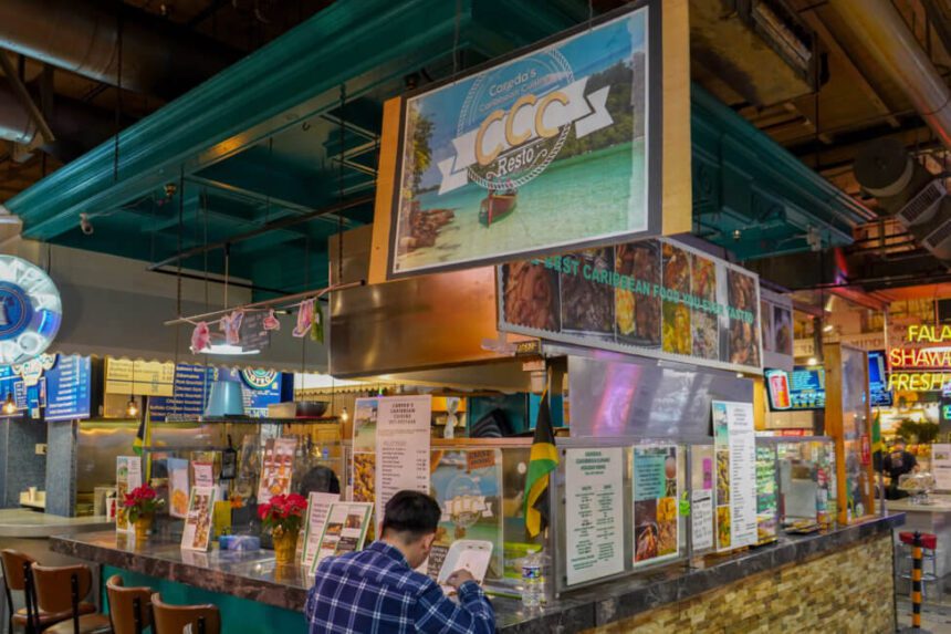 A sign with a beach as the background hangs above a counter. A man in a blue and white plaid shirt sits and eats at the counter.