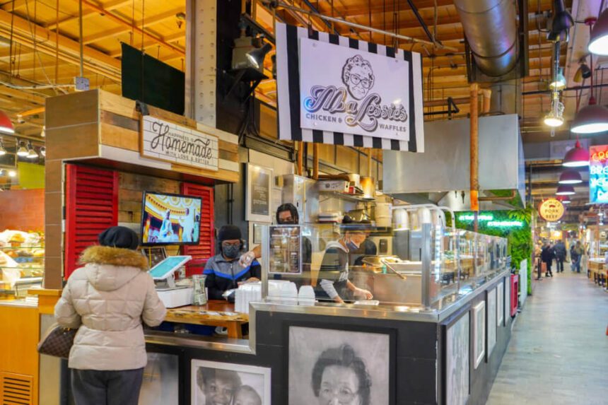 A stall is shown inside of Reading Terminal Market. A black and white sign with a woman's face drawn on it hangs above the stall and reads, "Ma Lessie's Chicken & Waffles." A woman in a winter coat stands off to the left in front of a register. There is another woman standing behind the register, helping the customer.