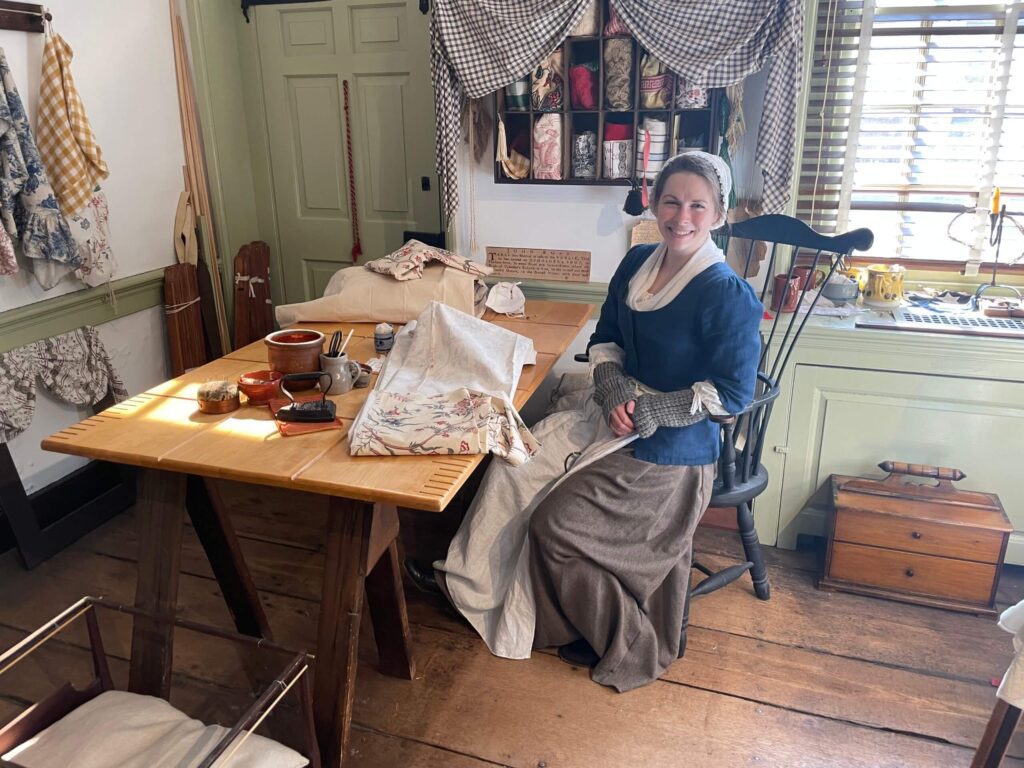 A woman portraying Betsy Ross sitting at a table inside a historic house. The woman looking at the camera and smiling.