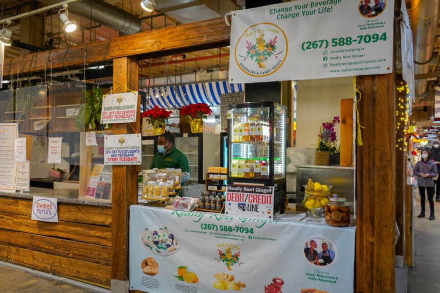 A wooden stall inside of Reading Terminal Market is shown. A man is standing behind the counter. The counter is filled with items and a fridge with drinks.