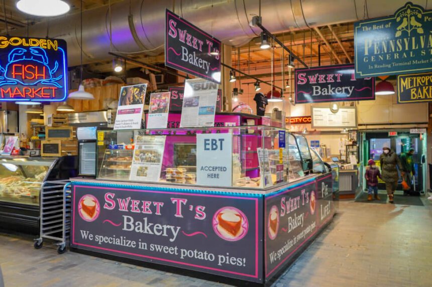A bakery station is shown inside of the Reading Terminal Market. The sign outside of the stall reads Sweet T's Bakery. There are pink and white signs.