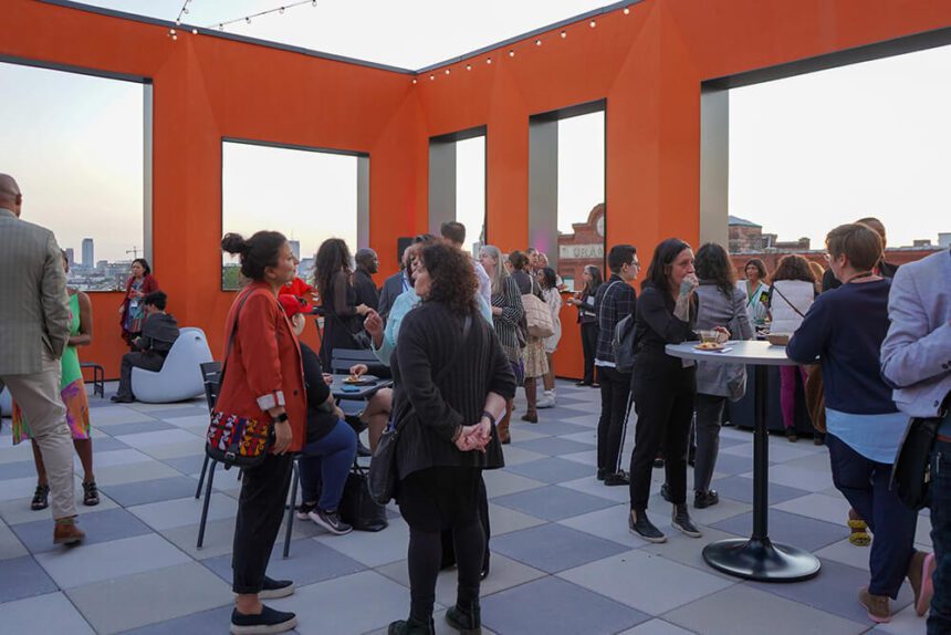 People are shown mingling on a rooftop. The walls surrounding them are painted a bright orange. Through the open windows, there are stunning views of the Philadelphia skyline.