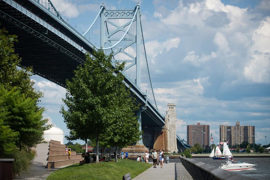 The Ben Franklin Bridge soars overhead under a bright blue sky. Boats are seen on the water. People are walking along a path. Children are seen standing and playing off to the left. Trees and green grass line the trail.