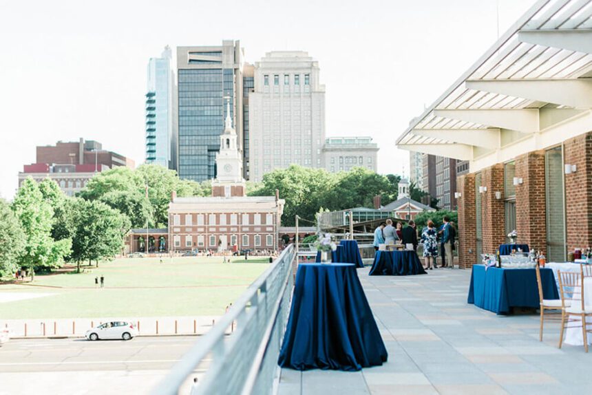 Independence Hall is off in the background. Multiple skyscrapers are behind it. The lush green lawn in front of Independence Hall is off to the left. There is a balcony overlooking the lawn set up with tables and chairs, high top tables covered in blue tablecloths, and a table set up with drinks on top of it. A group of people are shown socializing in the corner.