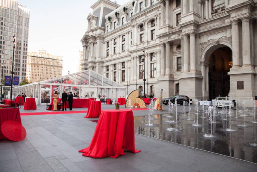 High top tables covered with red tablecloths are set up throughout Dilworth Park, an outdoor space next to Philadelphia City Hall. Fountains to the right spray water into the air. Off to the left in the background, there are three individuals standing in front of a white and clear tent. Two of the individuals are dressed in all black suits. The third individual is wearing a gold gown. Inside of the tent, there are red and black balloons hanging from the ceiling.