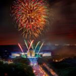 Fireworks light up the night sky above the Philadelphia Art Museum. The Benjamin Franklin Parkway is packed with people. The stage in front of the museum lights up with red, white, and blue lights.