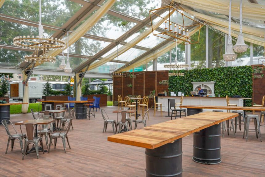 A large tent is set up outdoors. The roof is clear. There are trees outside of it. Inside of the tent, there are shorter tables set up with metal chairs around them. There is a taller table set up using a wooden slab as the counter and two barrels for the base. Back in the distance, a bar is set up in front of a green wall. Chandeliers hand from the roof of the tent.