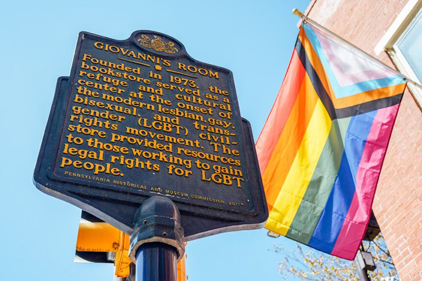 A blue historical marker for Giovanni's Room is shown. The writing on the marker is a bright yellow. A rainbow flag sways in the wind behind it to the right.