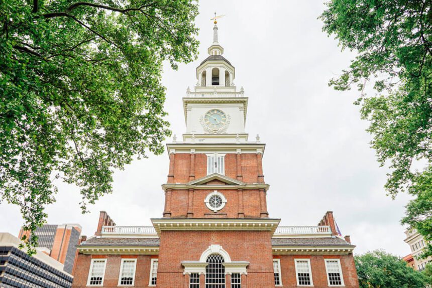 In the center is Independence Hall, a historical building. It is tall and made out of red bricks with white architectural details. Green trees hang on either side. The sky above is white. A clock is shown toward the top and in the middle of the building.