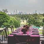 The Philadelphia skyline stretches across the shot. There are green trees in front of it. In the very front is a table covered in a burgundy tablecloth set for a dinner. There are wine glasses on the table. Each table setting has silverware and a cloth napkin. There are four chairs on either side of the table. Down the center of the table, there are beautiful flower arrangements.