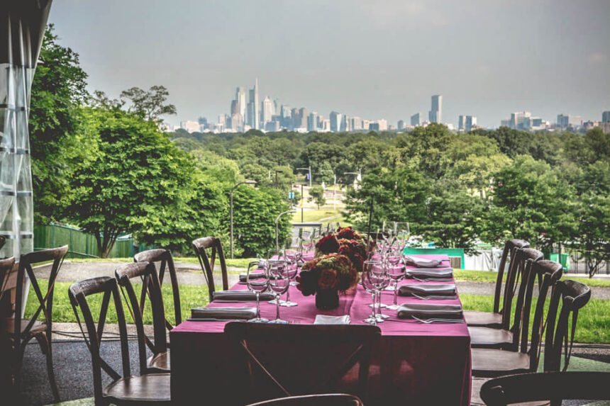 The Philadelphia skyline stretches across the shot. There are green trees in front of it. In the very front is a table covered in a burgundy tablecloth set for a dinner. There are wine glasses on the table. Each table setting has silverware and a cloth napkin. There are four chairs on either side of the table. Down the center of the table, there are beautiful flower arrangements.