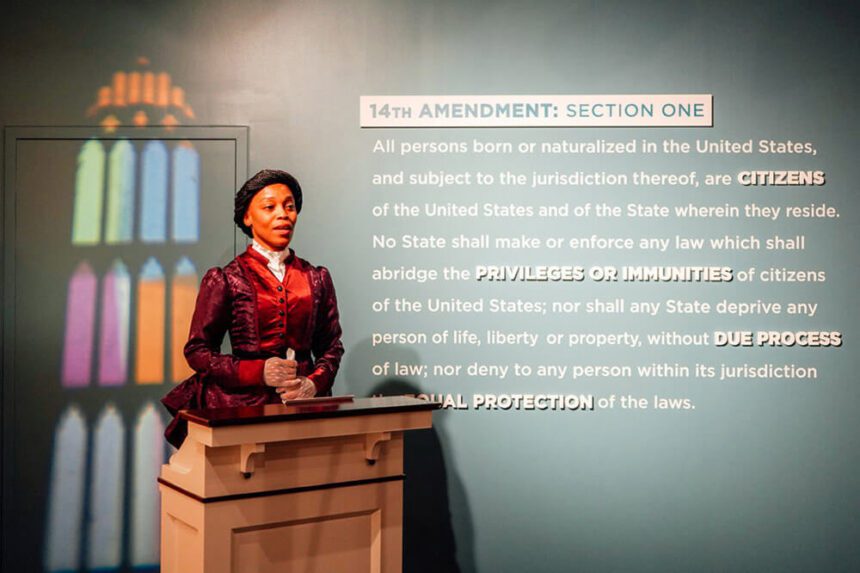 A woman in a maroon and red dress stands at a podium slightly off to the left. Beside her on the wall is the 14th Amendment written out in white writing against a gray backdrop.