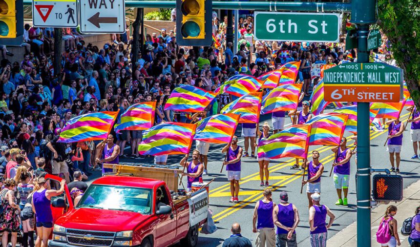 A group of men wearing purple tank tops march down the street waving rainbow flags as part of a Pride Parade in Philadelphia. Spectators watch from the sides.