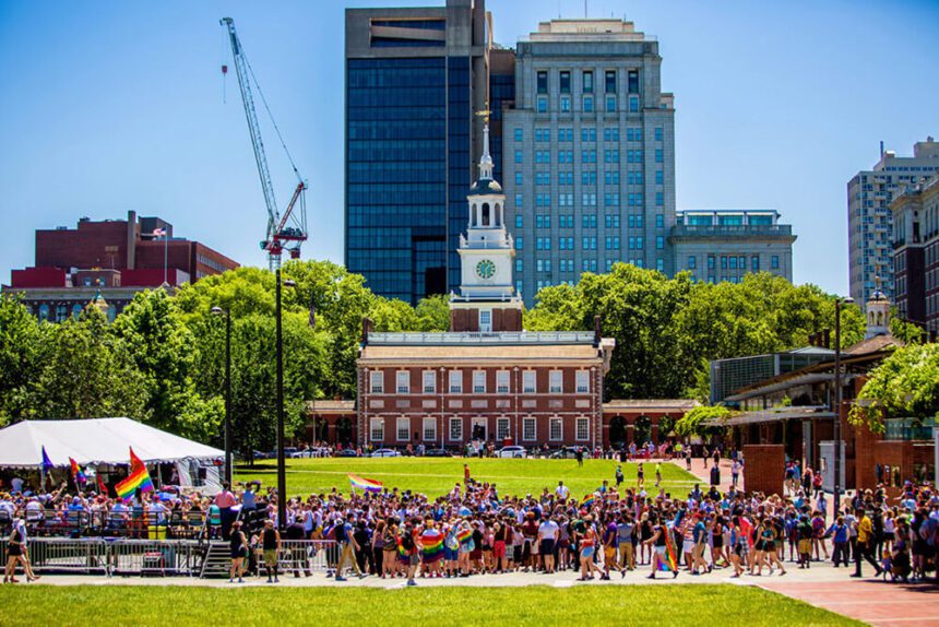 Independence Hall is in the background. A group of people are gathered in front. Some are shown with Pride rainbow flags. Others are dressed in bright colors. The grass is bright green. The sky overhead is a bright blue. 