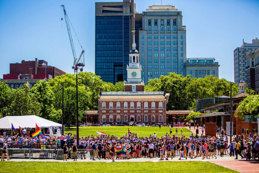 Independence Hall is in the background. A group of people are gathered in front. Some are shown with Pride rainbow flags. Others are dressed in bright colors. The grass is bright green. The sky overhead is a bright blue.