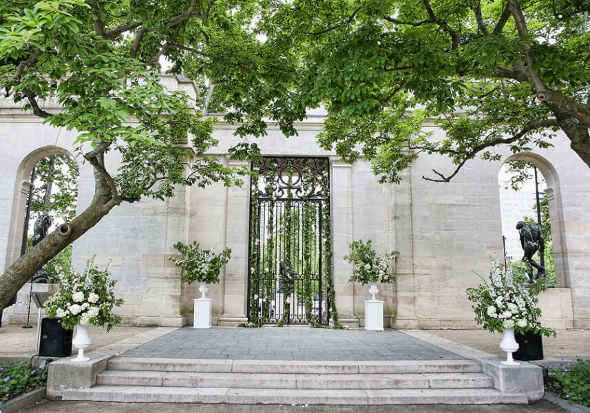 Large trees on either side provide shade over a walkway. The lush green leaves cover the top half of the shot. There are three concrete steps onto a landing in front of a gate. On either side of the gate, there is a planter with an elaborate display of flowers. Similar planters are placed on wither side of the set of steps. There are statues on either side off toward the back under arches.