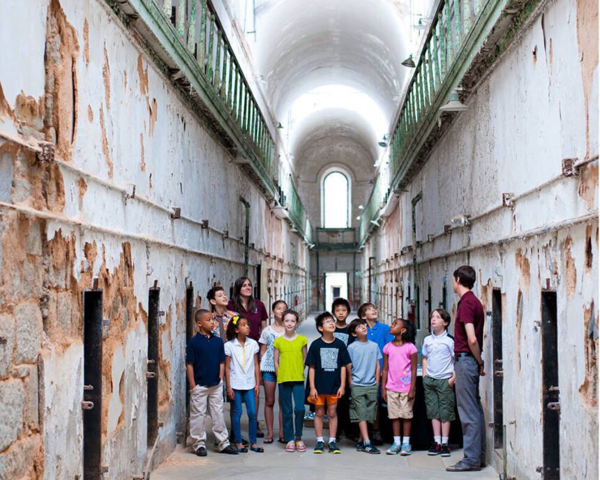 A group of young children stand in a hallway in Eastern State Penitentiary, accompanied by Eastern State Penitentiary employees giving the students a tour of the site. Crumbling cell blocks surround them. The children are shown looking around as they listen.