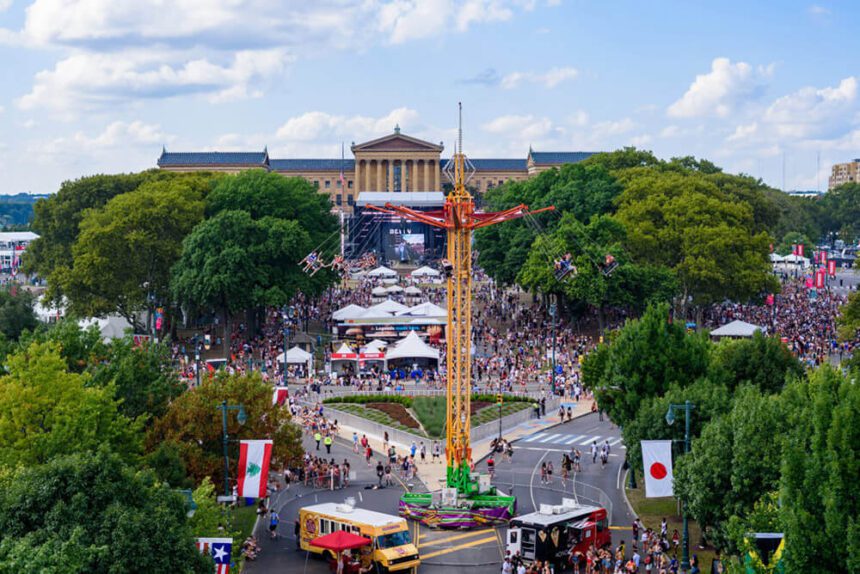 A tall yellow amusement ride is in the center with people in swings hanging from it. The Philadelphia Museum of Art is in the distance. Below the ride, people are shown walking and partying on the Benjamin Franklin Parkway. There are crowds of people wearing red, white, and blue by white tents set up throughout the space.