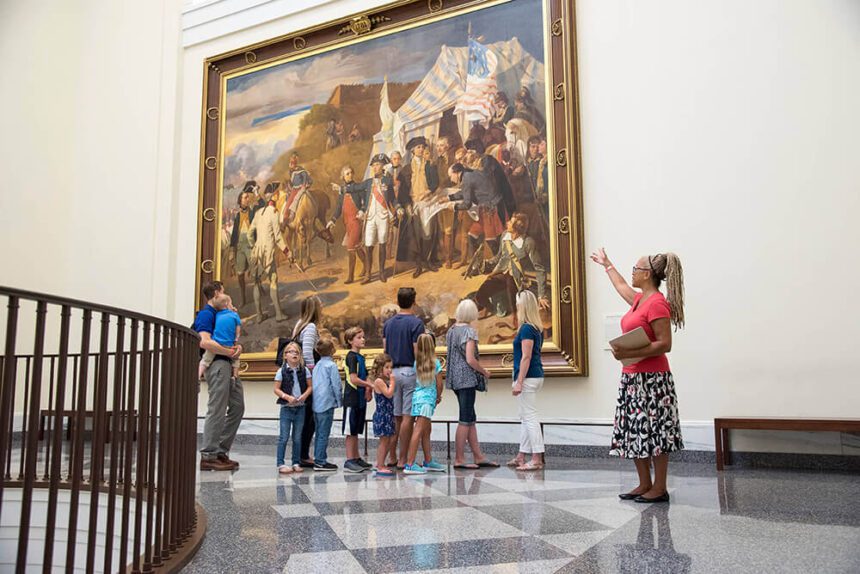 A tour guide stands off to the right, motioning with her arm toward a massive painting hanging on the wall. A group of people stand in front of the painting, admiring it. The group consists of both adults and children. The painting shows a historical moment during a war.