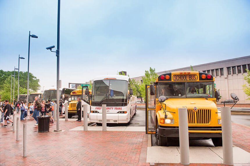 A yellow school bus is shown off to the right, a white shuttle bus is to the left of it. The sky overhead is clear. People are shown off to the left walking away from the buses.