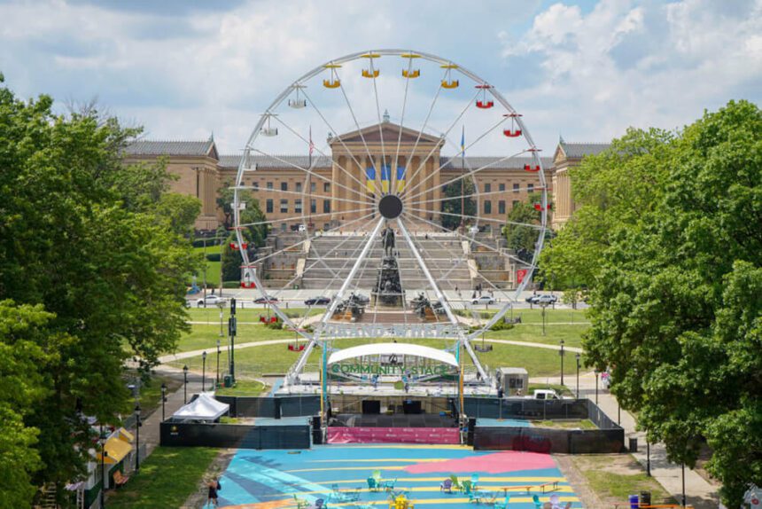 A giant white Ferris Wheel is shown in the center. Its cars are colorful, some yellow, some red. The Philadelphia Museum of Art is behind it. In front of the Ferris Wheel is a colorful mural with bright chairs set up.
