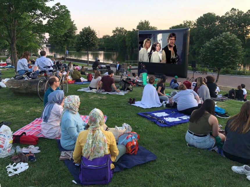 People are shown sitting on picnic blankets on the grass, looking up at a large movie screen set up in a park. Trees are shown throughout. 