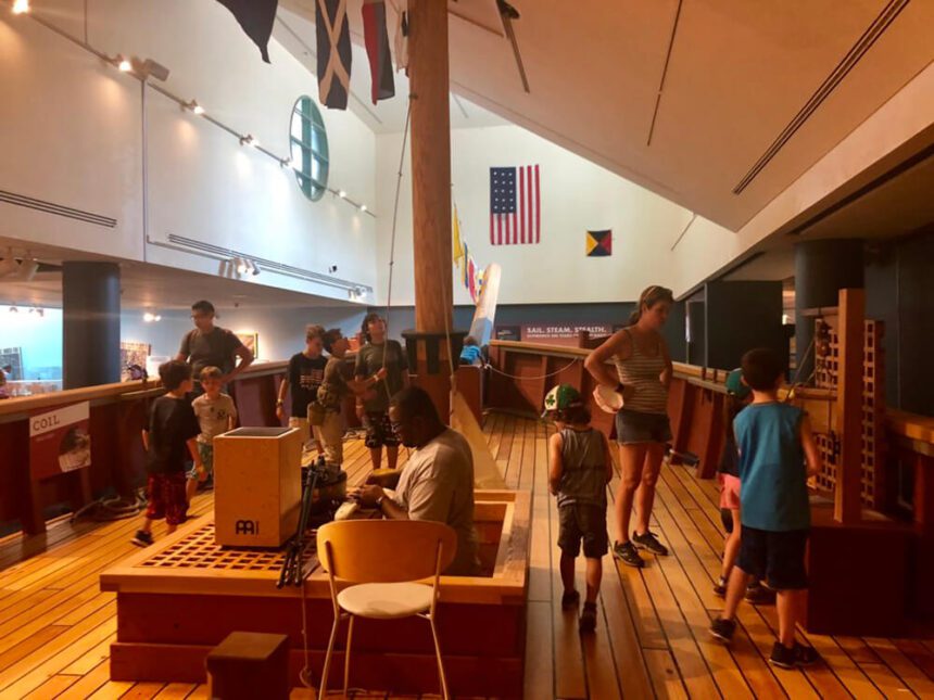 There are multiple young children and adults gathered in a space made to resemble a ship. There are interactive elements shown. An American flag hangs on the wall behind them.