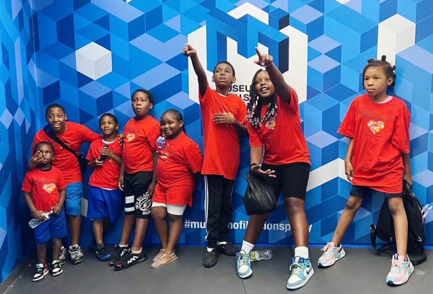 Eight young children wearing bright red t-shirts are shown standing up against a blue and white wall inside of a museum. Some of the children are pointing, others are looking up.