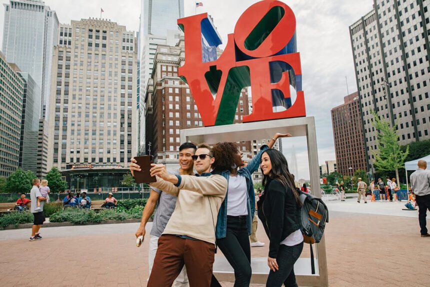 A group of four individuals are shown posing for a selfie in front of the iconic red LOVE statue. There are buildings in the background. There are people shown in the distance.
