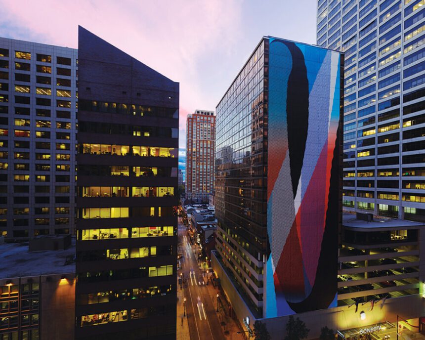 Skyscrapers are shown. To the right, a skyscraper hotel is covered with a colorful mural of reds, blues, and black. The street below is lit up by streetlights. The building to the left of the hotel has rooms lit up in a yellow hue. The sky above is a light blue and pink.