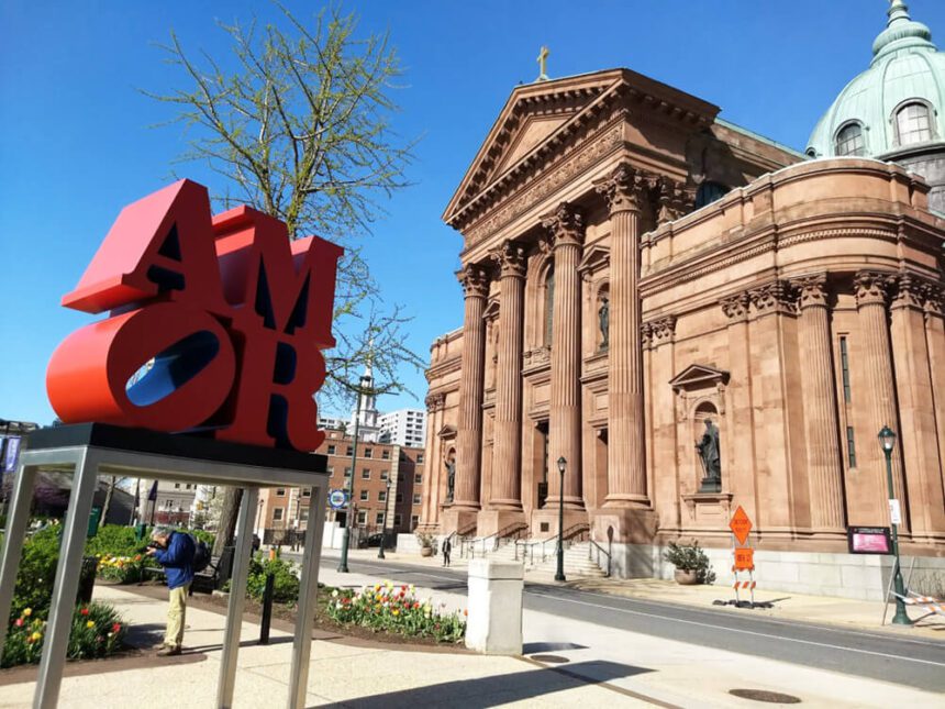A large red sculpture is shown on a silver pedestal to the left. The red letters spell AMOR. To the right, there is a large church. It is a light brown with a mint green dome and gold crucifix on top. The sky above is a clear, bright blue.