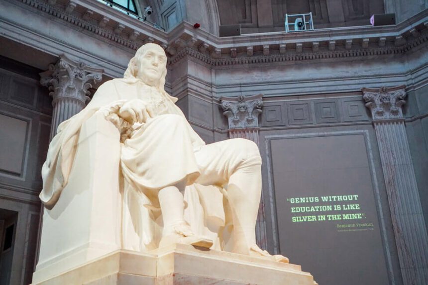 A large white statue of Benjamin Franklin is shown. He appears to be sitting in a chair looking off into the distance. There is a quote projected on the wall behind the statue that reads, "Genius without education is like silver in the mine."
