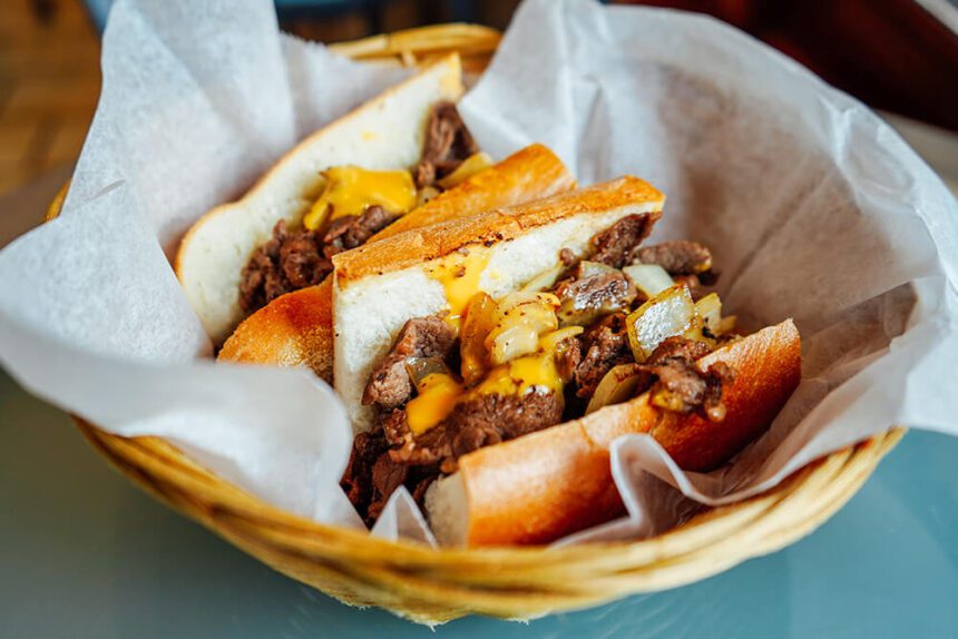 A sandwich is shown. The roll is a light brown. The meat inside is like an almost dark gray. The meat is smothered in yellow cheese. The sandwich is in white paper in a wicker basket.