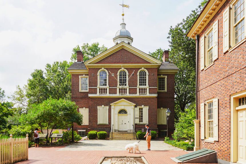 A large brick building is shown. In front of it, there is a man walking a white dog. There are lush green trees and bushes on either side of the building. The sky above is a bright, light gray.