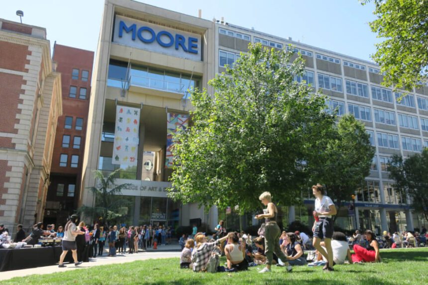 A large group of people are shown sitting on a green lawn in front of a large building that has MOORE written in large blue letters across the top of it. There are more students gathered by the entrance to the school. The sky above is a clear blue.