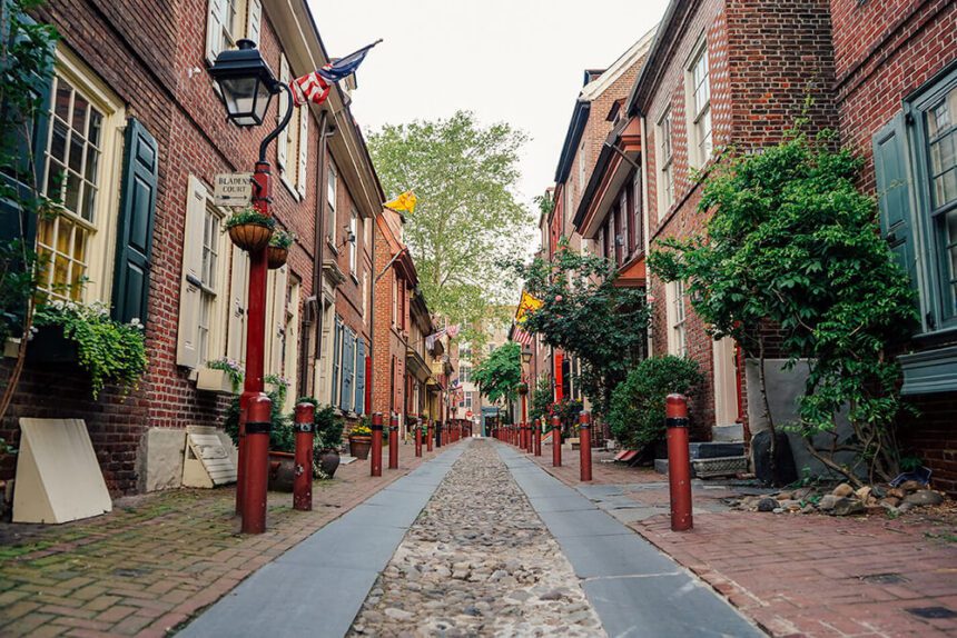 A cobblestone street is shown. There are brick buildings on either side of the street. There are green trees scattered throughout the outdoor space. 