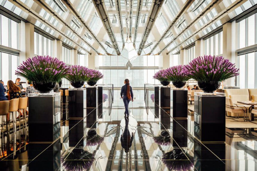 Six large floral arrangements line a tiled area inside of the Four Seasons Hotel. A woman walks in the center, she is dressed in navy blue and has long red hair. There is natural light coming in from the windows and the ceiling. There are chairs off to the right. Off to the left, there are people sitting down at a bar.