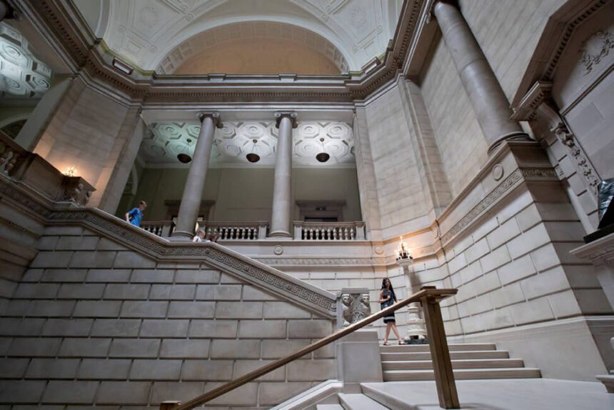 A large staircase is shown. A woman appears to be walking down the staircase. A few more individuals appear to be also walking down the steps. There are two large columns shown. The indoor space has a very large ceiling.